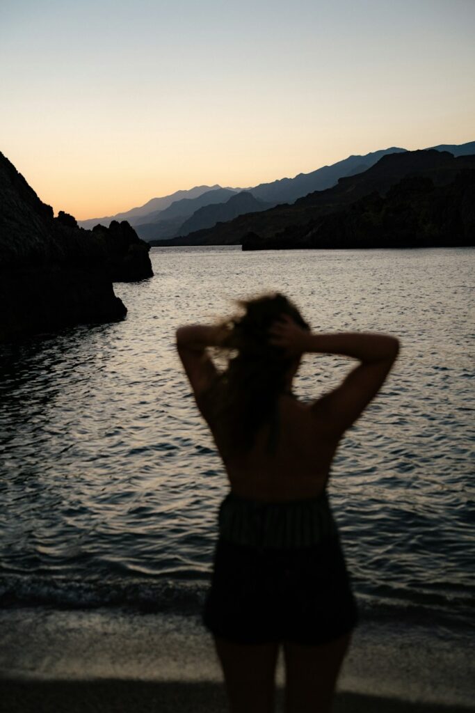 A person in silhouette with hands in hair facing a calm sea and mountains